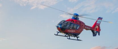 A red and blue Devon Air Ambulance helicopter flying with a blue sky, slightly cloudy background